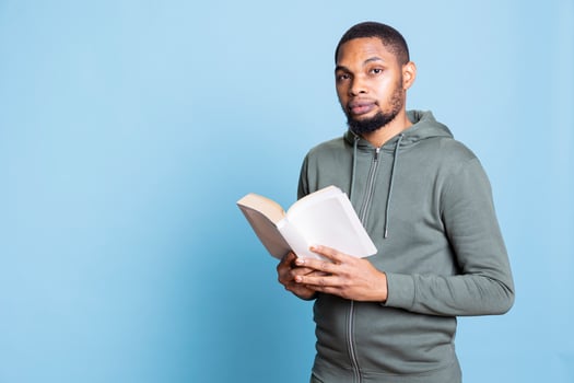 African american cheerful guy enjoys reading a book in studio, feeling happy with literature and novel against blue background. Confident book lover analyzing the synopsis and characters. photo