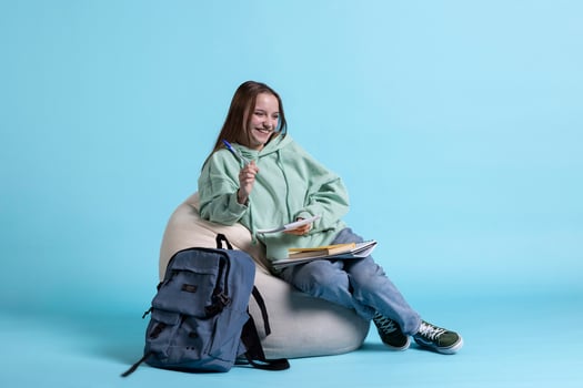 Portrait of jolly girl laughing while sitting on beanbag and writing on notebook, studio background. Overjoyed schoolgirl with notepad in arms feeling amused while preparing homework photo