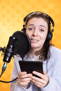 Portrait of smiling woman reading ebook from ereader, recording audiobook using microphone, studio background. Jolly narrator using mic to produce digital recording of novel, reading text from tablet photo