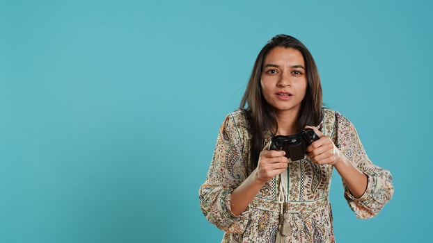 Cheerful indian gamer showing thumbs up sign gesturing while holding controller after winning videogame. Joyful woman enjoying games, doing approving hand gesture, studio background, camera A photo