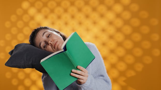 Woman struggling to keep eyes open while reading book for school exam, feeling exhausted, studio background. Tired student feeling sleepy after doing homework, putting head on pillow, camera B photo