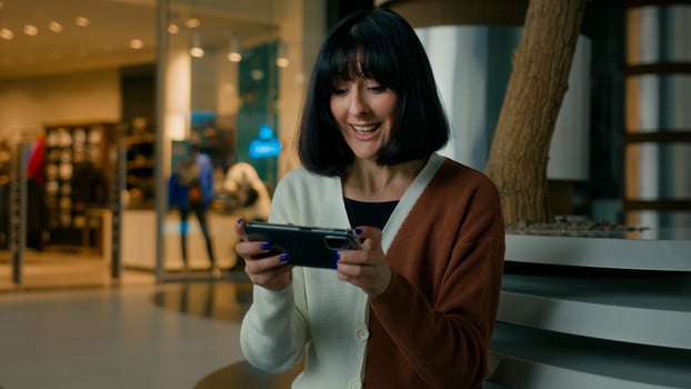Woman lady female gamer girl indoors in shopping mall sitting playing with mobile phone in online game in net smartphone gaming app emotional enjoy play competition win celebrate victory winning photo