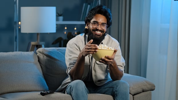 Bearded man in glasses indian male bachelor after work sits on sofa in dark living room turns on TV with remote control choosing channel watching online movie film program eating popcorn photo