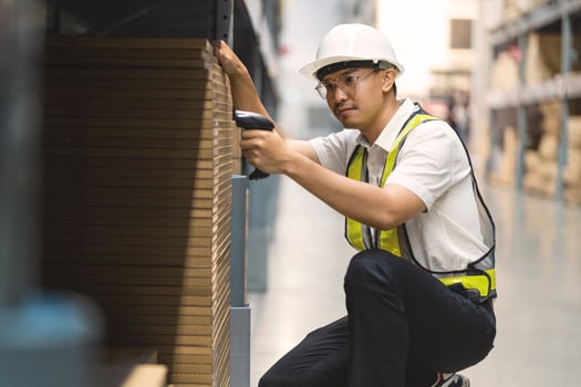 A man in a safety vest is sitting on the floor in a warehouse. He is holding a barcode scanner and looking at a box photo