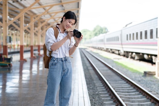 Young Asian woman holds a camera and takes photos of a train while waiting for the train at the train station to travel.