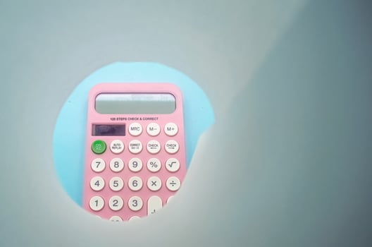 A pink calculator with white buttons is viewed through a circular cutout. The calculator is placed on a light blue background, creating a unique and creative composition photo
