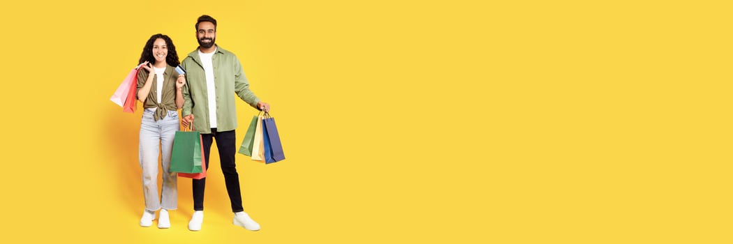 Shopping. Smiling middle eastern couple with credit card and shopper bags posing, bought a lot of purchases during seasonal sale over yellow background in studio. Full length shot photo