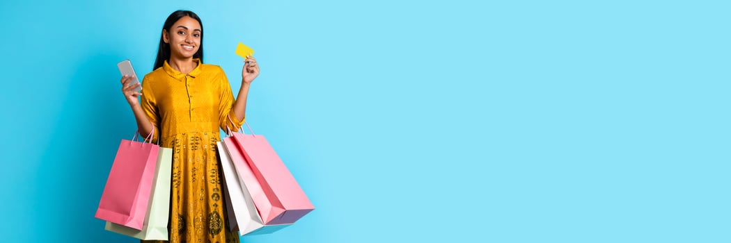 Shopaholic. Happy Indian Customer Woman Holding Smartphone, Credit Card And Shopping Bags, Making Purchases Via Application On Mobile Phone While Standing Over Blue Studio Background photo