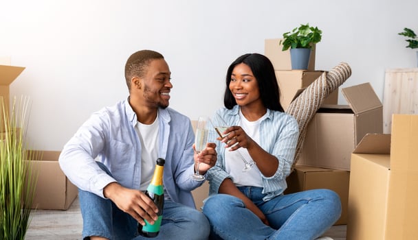 Young african american couple celebrating moving to new apartment, sitting among cardboard boxes and drinking champagne. Home owners and family housing concept photo