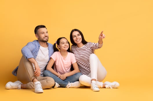 A loving family of three shares a joyful moment against a bright yellow background. The parents sit closely with their daughter, who points at copy space while they all smile photo