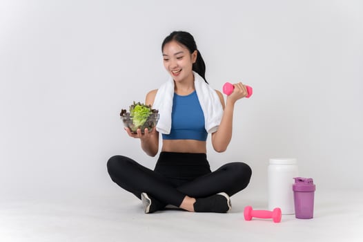 A happy, healthy Asian woman sits on a white studio background, holding a dumbbell and a salad bowl. photo