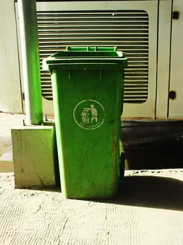 A green trash can with a sticker on it, sits empty on the ground. The trash can is located next to a metal pole and a building photo