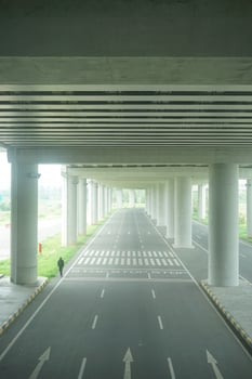 A long, straight road passing through a concrete overpass structure with multiple arched openings, surrounded by green foliage at new yogyakarta international airport photo