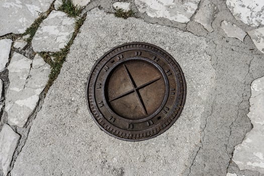 A round metal manhole cover with a cross design in the center, embedded in a cracked concrete surface with some grass growing between the cracks photo