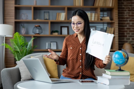 Asian woman conducting online lesson from home. She sits on sofa with laptop, holding a whiteboard marked Lesson, engaged in interactive teaching session, conveying education and technology. photo