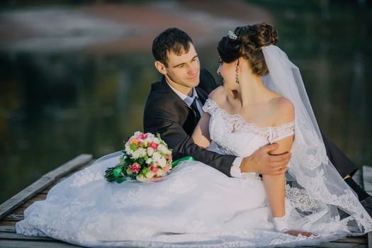 A bride and groom are sitting on a dock, with the bride holding a bouquet photo