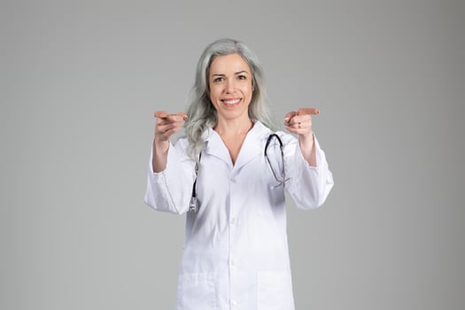 A mature woman in a white lab coat stands confidently, smiling while pointing toward the viewer. She has long gray hair and a stethoscope around her neck, indicating her role in healthcare. photo