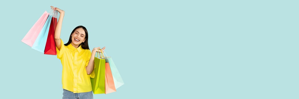 Happy customer. Emotional young brunette woman holding bright shopping bags and smiling, raising hands up, looking at camera over blue background, copy space photo