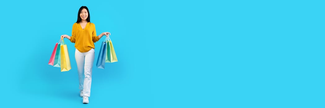 Pretty young Asian woman with colorful shopping bags walking over blue studio background, panorama with copy space. Season sale concept photo