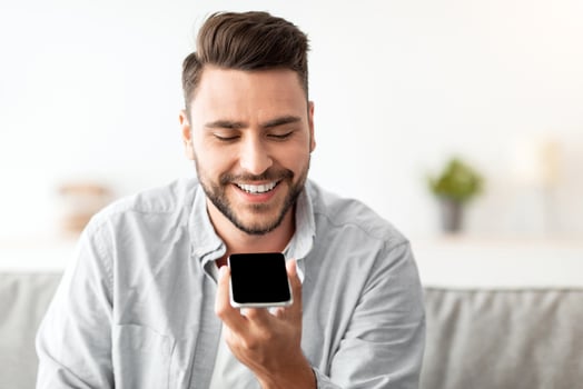 Happy handsome man recording voice message on smartphone or using virtual assistant while resting on sofa at home. Smiling guy enjoying smart technologies for communication, free space photo