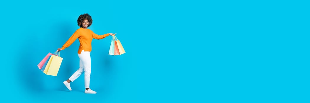 Positive carefree beautiful young black woman in stylish outfit shopaholic walking with purchases colorful shopping bags over blue studio background, smiling at camera, copy space, full length photo