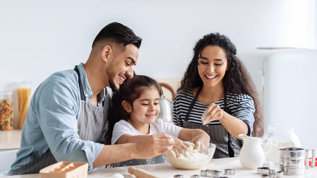Cheerful millennial arab parents making cookies with their little daughter at kitchen, blending dough ingredients in glass bowl and laughing, father, mother and kid having fun while baking at home photo