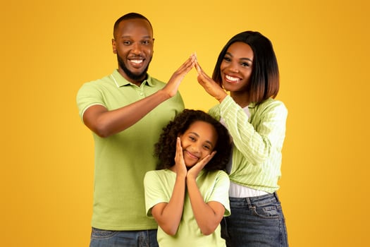 Family care, protection and insurance concept. Young black parents making symbolic roof of hands above little daughter while standing over yellow background, studio shot photo