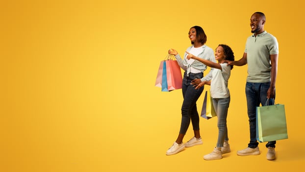 Family shopping. Pretty girl pointing finger at free space, showing something to parents, standing together with shopper bags over yellow background, panorama with empty space photo