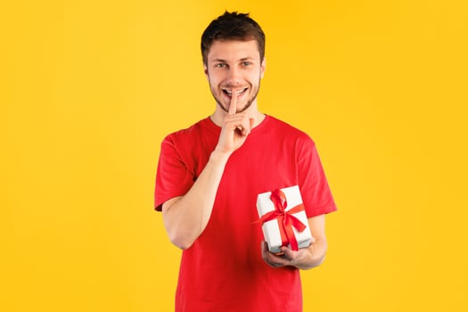 Surprise Gift. Portrait of smiling young guy holding wrapped present box and showing silence sign, isolated over blue studio background, greeting and celebrating special day or event photo
