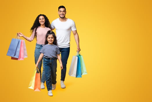 Shopping Day. Cheerful Young Middle Eastern Family Of Three Walking With Bright Shopper Bags Over Yellow Background In Studio, Happy Arab Parents And Female Child Enjoying Seasonal Sales, Copy Space photo