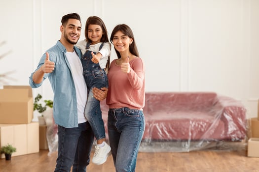 Moving Day. Portrait of cheerful daddy, mum and daughter posing in new apartment, standing in empty living room showing thumbs up sign gesture. Cheerful parents holding girl on hands, free copy space photo