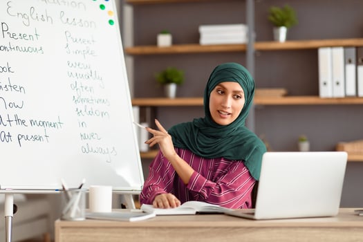 Modern Distance Learning. Muslim English teacher making conference chat with students using pc laptop. Lady in hijab sitting at desk pointing on board teaching from home during covid outbreak photo