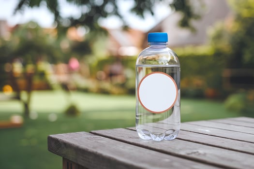 A blank white circular sticker mockup is attached to a water bottle which is placed on a table in garden photo