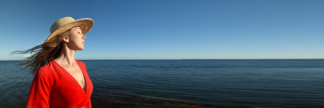 Panoramic Portrait profile of a young 30-year-old European woman in a straw hat and a red dress. Looking away with her eyes closed against the background of a clear sky and blue sea. photo