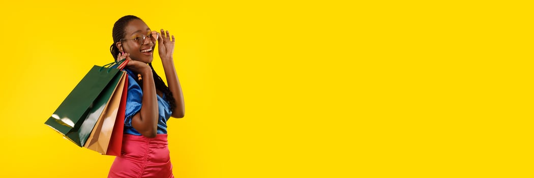 Pretty young black woman with colorful shopping bags posing over yellow studio background, panorama with copy space. Season sale concept photo