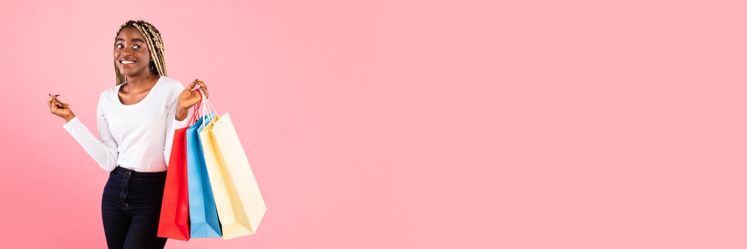 Happy young African American woman with braided hair enjoying shopping during sale season, holding purchases over pink studio background, copy space photo