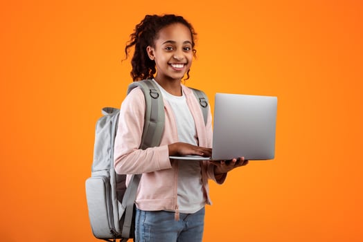 Portrait of smiling African American girl holding and using laptop computer, wearing backpack, typing on keyboard, posing and looking at camera standing isolated over yellow studio background photo