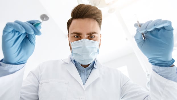 Male dentist in protective face mask and medical gloves holding dental tools, making checkup for patient. Young man stomatologist doing treatment, closeup portrait, low angle shot. photo