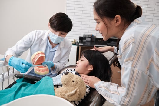 Asian male pediatric dentist demonstrates teeth brushing to girl with toothbrush and dentures in dental clinic, her mother is nearby, and professional orthodontic healthcare worker in kid hospital. photo