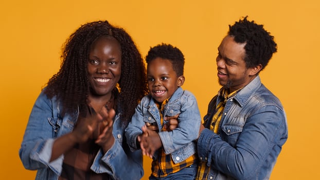 African american young couple clapping hands and dancing with their small kid against yellow background. Mother and father feeling happy together with their young toddler. Camera A. photo