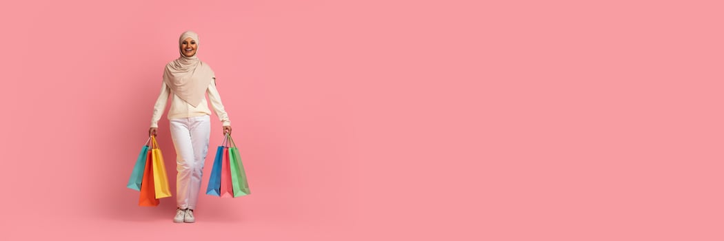 Cheerful young muslim woman walking with shopping bags over pink studio background, panorama with copy space photo