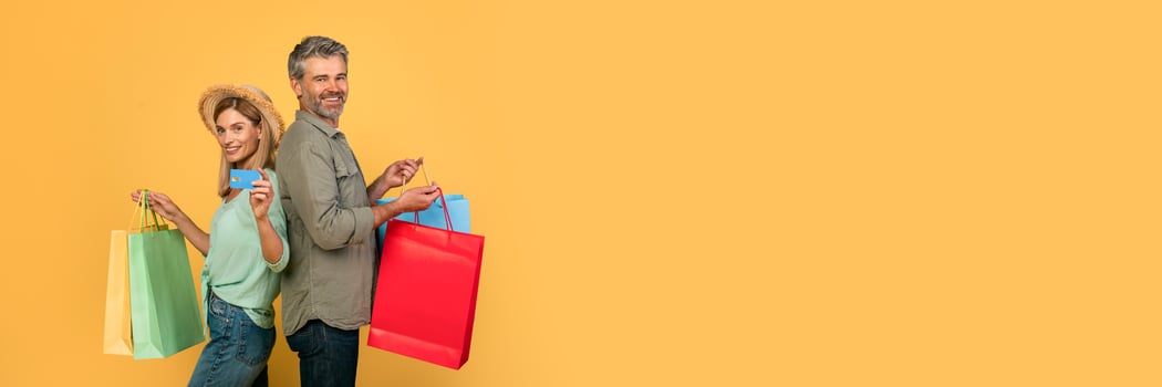 Portrait of middle aged caucasian spouses showing credit card and holding colorful shopper bags after purchases in shopping mall, standing over yellow background, studio shot photo