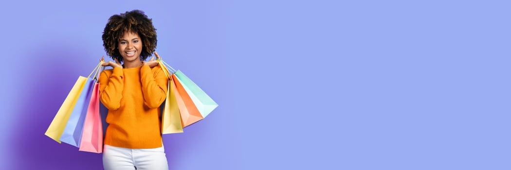 Shopping, retail. Positive carefree beautiful young black woman in stylish outfit shopaholic holding purchases colorful shopping bags over purple studio background, smiling at camera, copy space photo