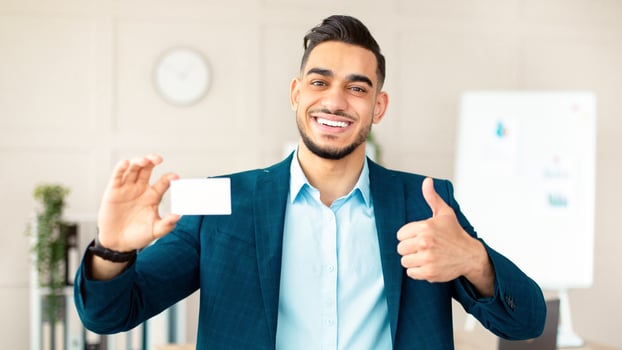 Happy Arab guy entrepreneur holding empty business card, showing thumb up gesture at company office, mockup for design. Smiling CEO manager demonstrating blank advertising card photo