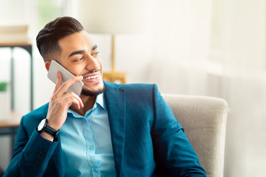 Joyful Arab entrepreneur in business suit talking on cellphone at office, sitting in armchair, having conversation with friend or customer. Office worker in formal wear speaking on smartphone indoors photo
