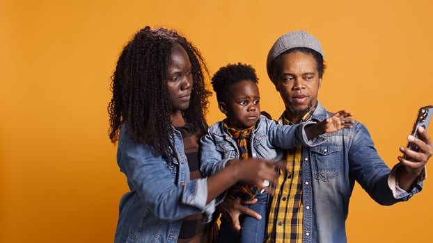 Young family smiling for selfies on smartphone with their boy against yellow background, capturing sweet memories together. Proud african american mom and dad posing with the child. Camera B. photo