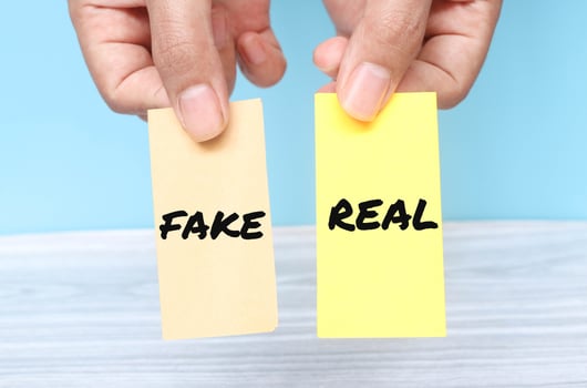 Hand holding the words Fake or Real on yellow shaped paper isolated on blue background. Real or fake business concept photo