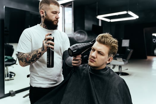 Barber makes hair styling with hair spray after haircut at the barber shop. Young handsome Caucasian man getting a haircut in a modern hairsalon. photo
