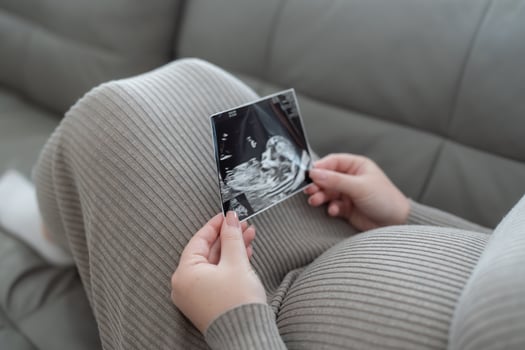 Gen Z pregnant woman holding ultrasound scan photo of a fetus sitting on a sofa. Concept of pregnancy, Maternity prenatal care