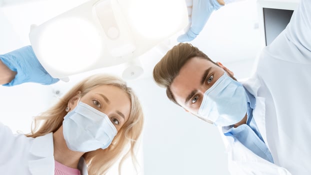Man and woman two dentists in medical face masks and rubber gloves doing treatment, turning on lamp, shot from dental chair. Modern stomatology, high class dental clinic concept, panorama photo
